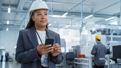 Portrait of a Black Female Engineer in Hard Hat Standing and Using a Smartphone at Electronics Manufacturing Factory. Technician is Writing a Message and Checking Her Schedule.