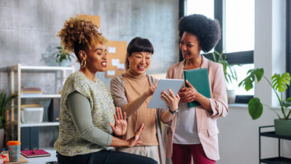 Three young business women talking about project while one holds tablet.