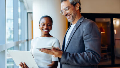 Two smiling business colleagues engaging in discussion while holding a laptop and tablet indoors. They appear in high spirits as they interact in a brightly lit setting.