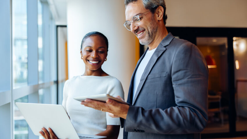 Two smiling business colleagues engaging in discussion while holding a laptop and tablet indoors. They appear in high spirits as they interact in a brightly lit setting.