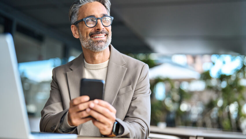 Happy middle aged business man using phone sitting outside office looking away.
