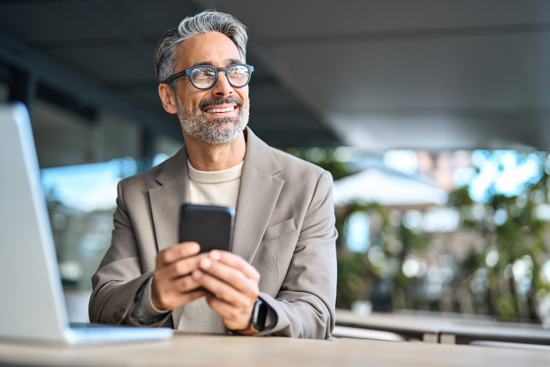 Happy middle aged business man using phone sitting outside office looking away.